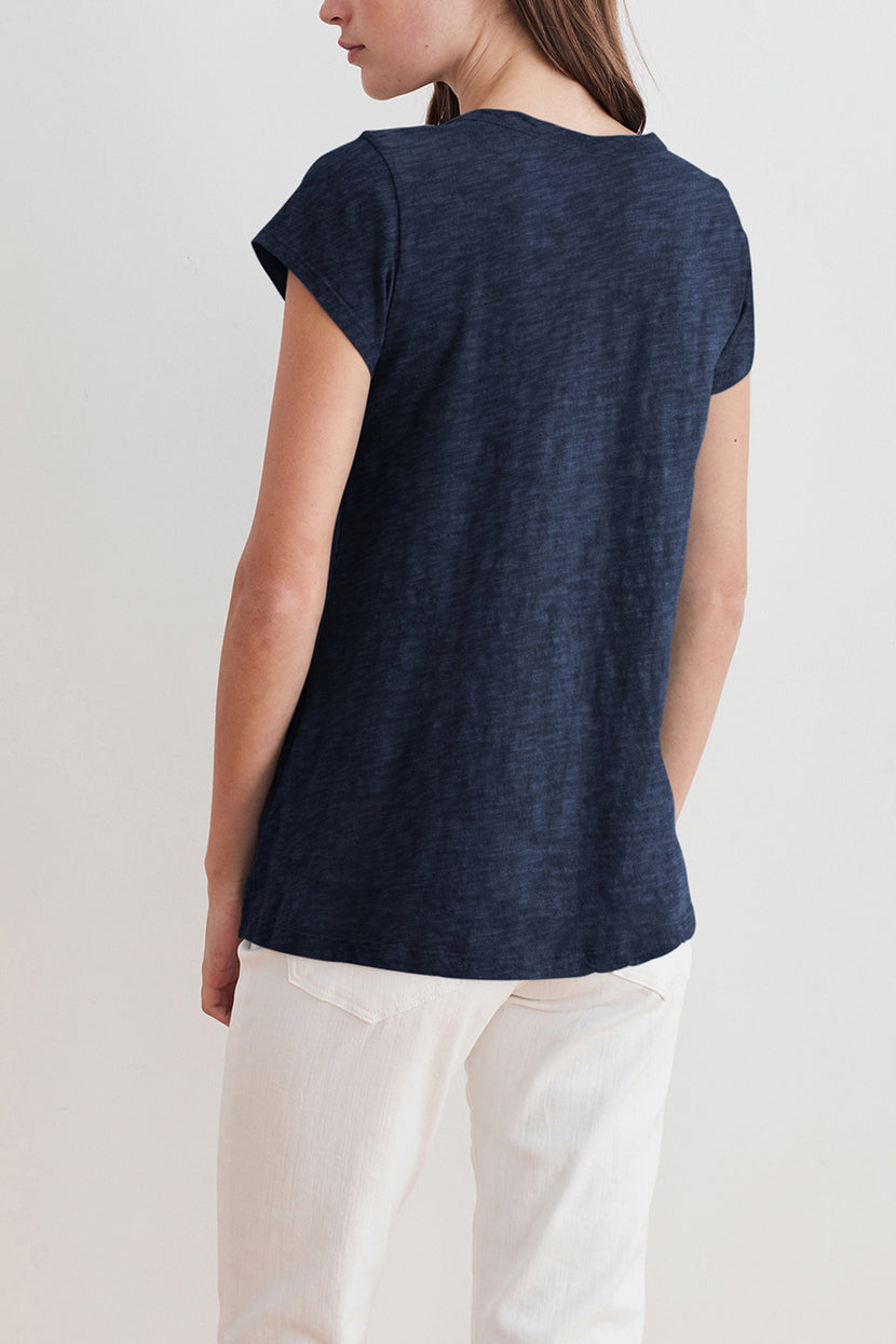 A person stands with their back to the camera, wearing a navy blue KIRA TEE by Velvet by Graham & Spencer made of cotton slub fabric and white pants, perfect for everyday wear.