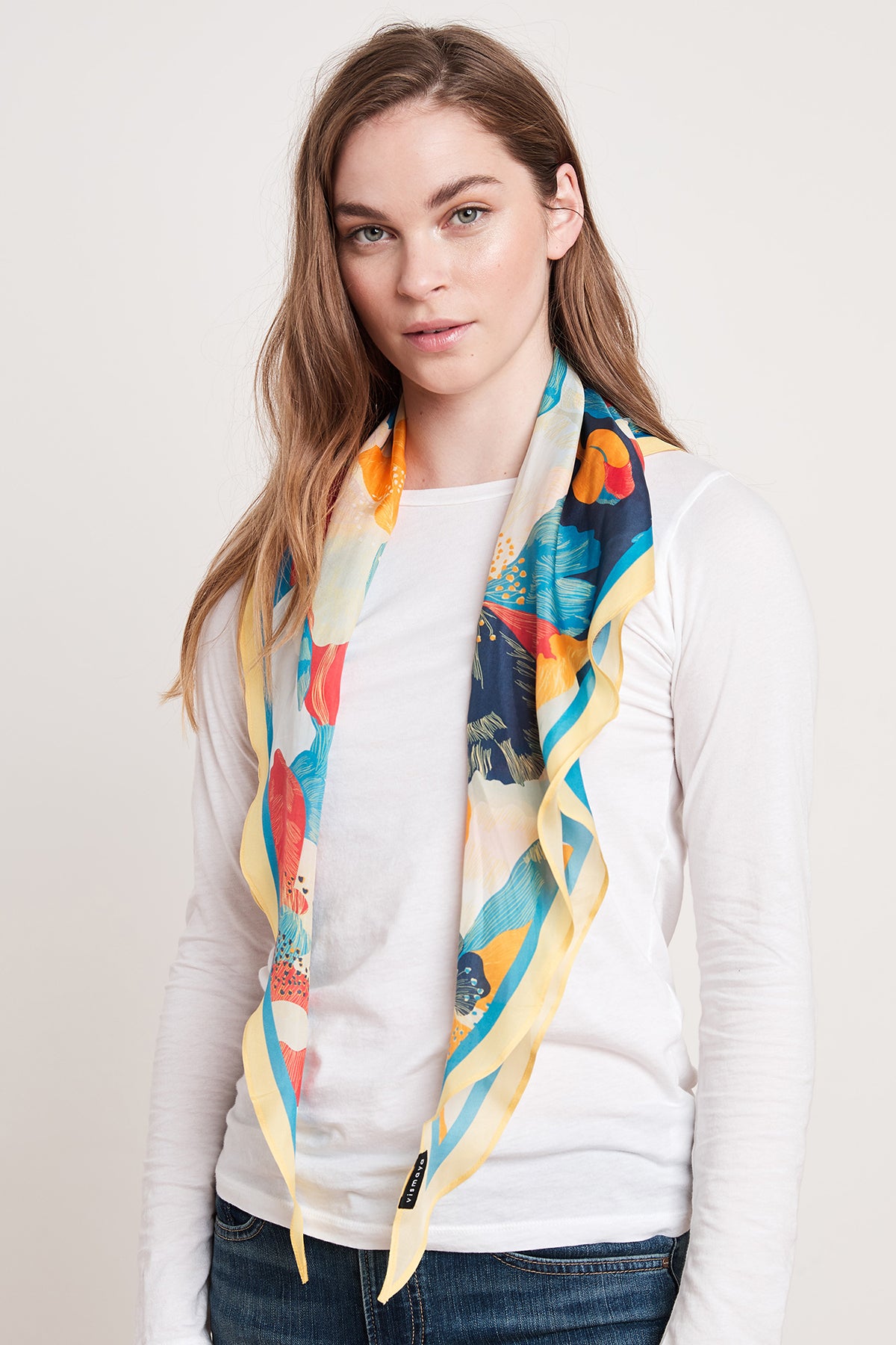   A person with long hair wears a white long-sleeved shirt and the DIAMOND SHAPE SILK SCARF by Velvet by Graham & Spencer, featuring blue, yellow, and red patterns, standing against a plain background. 