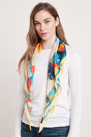 A person with long hair wears a white long-sleeved shirt and the DIAMOND SHAPE SILK SCARF by Velvet by Graham & Spencer, featuring blue, yellow, and red patterns, standing against a plain background.