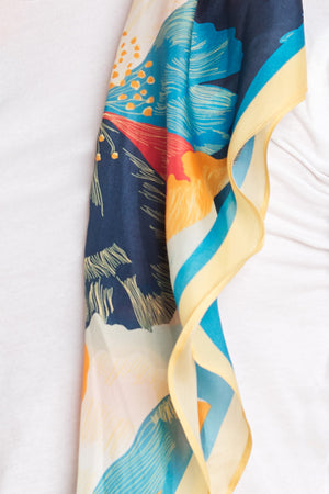 A close-up of a person wearing a white top with the DIAMOND SHAPE SILK SCARF by Velvet by Graham & Spencer featuring a colorful floral pattern in shades of blue, yellow, and red draped over their shoulder.