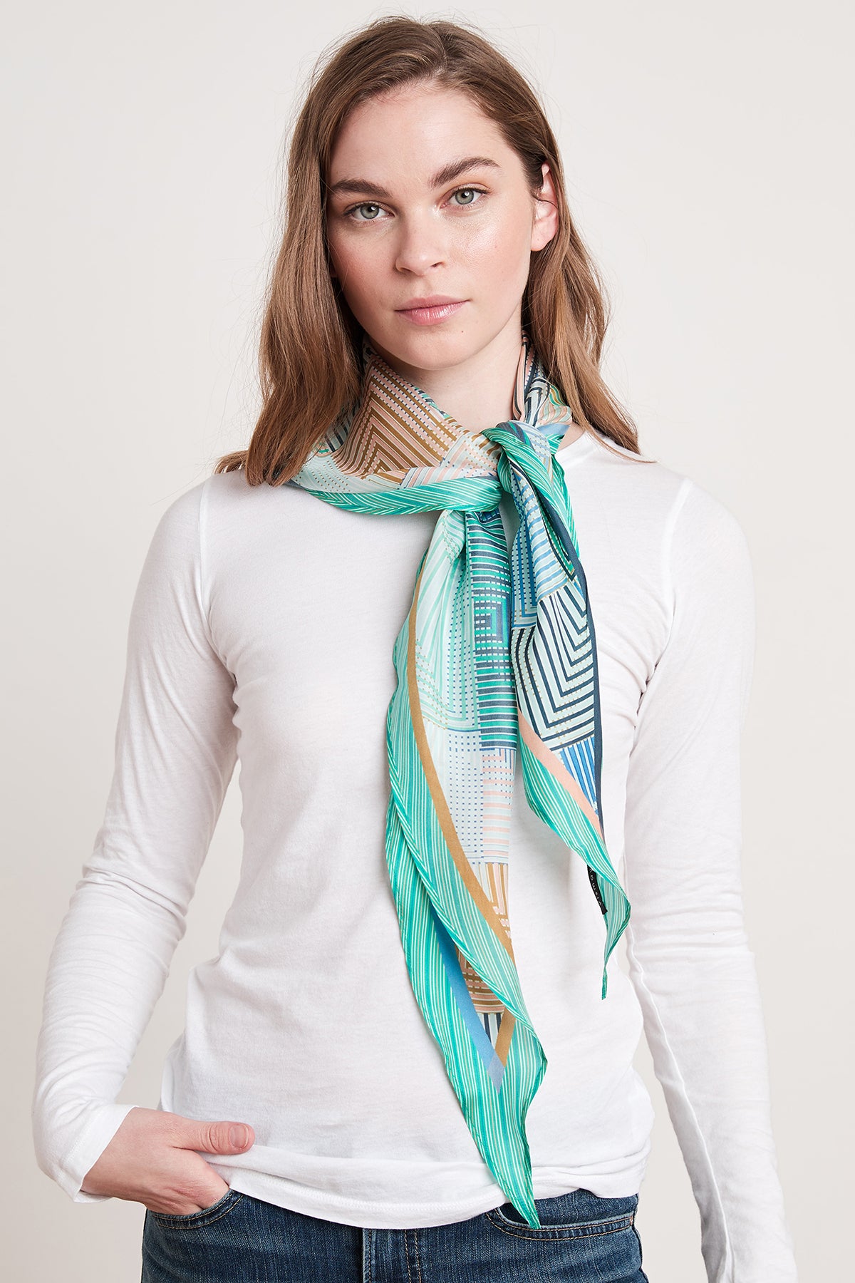   A woman with shoulder-length hair is wearing a white long-sleeve shirt and the Diamond Shape Silk Scarf by Velvet by Graham & Spencer in a green and blue pattern around her neck. She stands against a plain background with one hand in her pocket, presenting a relaxed diamond silhouette. 