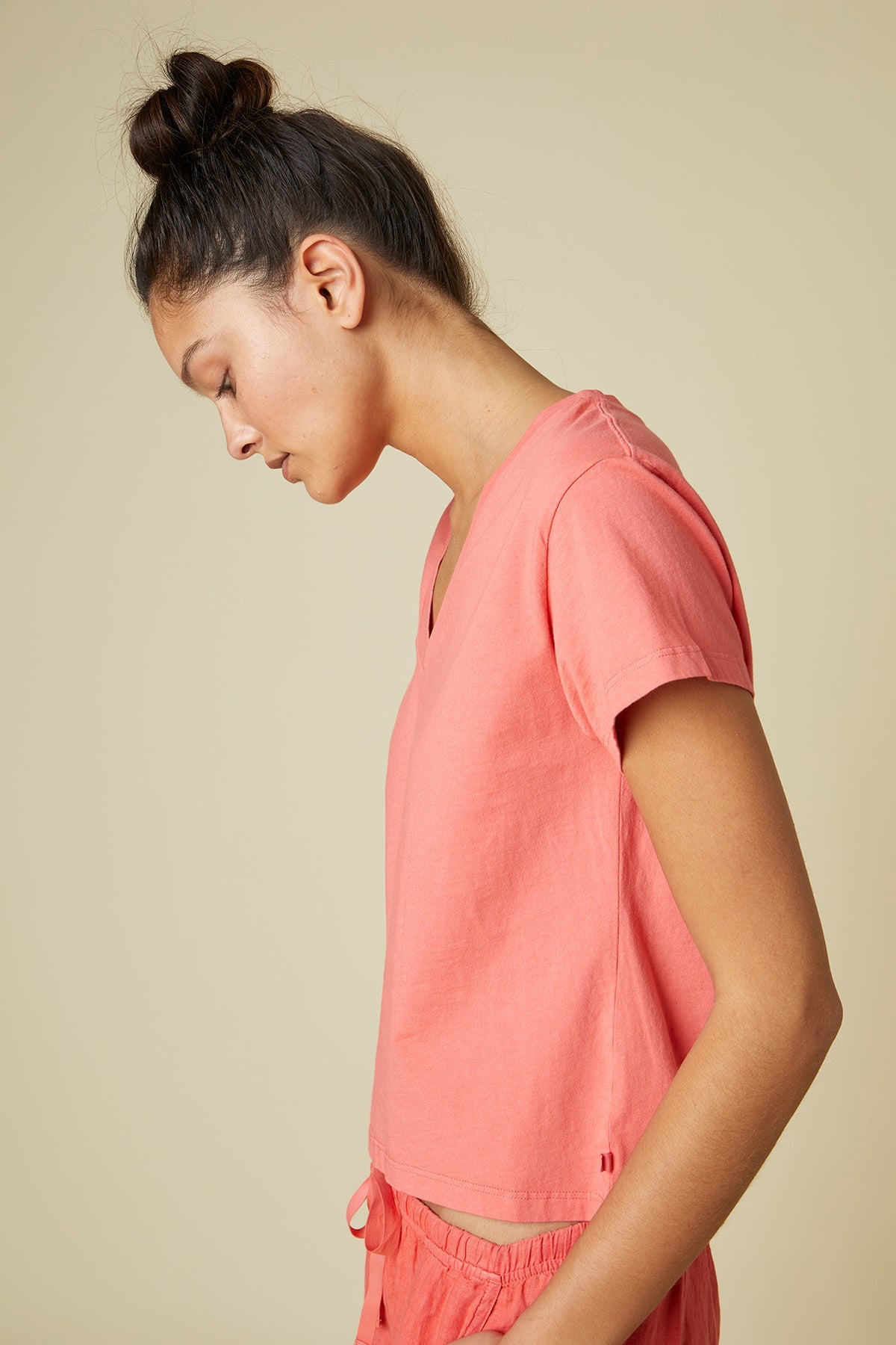   A person with tied-back hair wearing a pink Velvet by Jenny Graham VENICE TEE and matching bottoms made of organic cotton, looking downward with a neutral expression, is photographed against a plain beige background. 