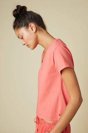 A person with tied-back hair wearing a pink Velvet by Jenny Graham VENICE TEE and matching bottoms made of organic cotton, looking downward with a neutral expression, is photographed against a plain beige background.
