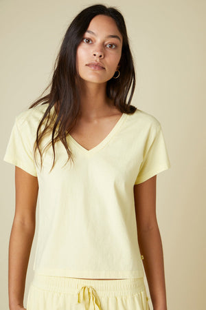 Woman with long dark hair wearing a light yellow VENICE TEE and matching drawstring pants made from organic cotton by Velvet by Jenny Graham stands against a beige background.