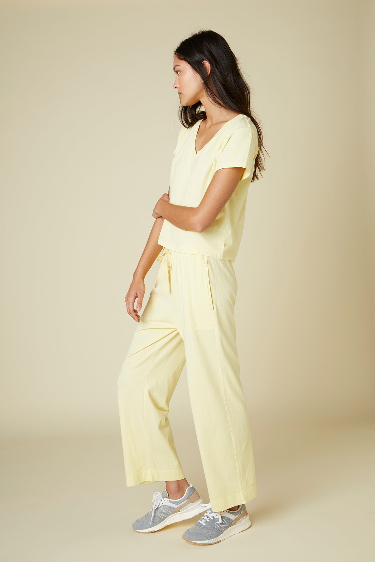 A woman stands in profile wearing a light yellow VENICE TEE by Velvet by Jenny Graham made from organic cotton and gray sneakers.-24063663997121