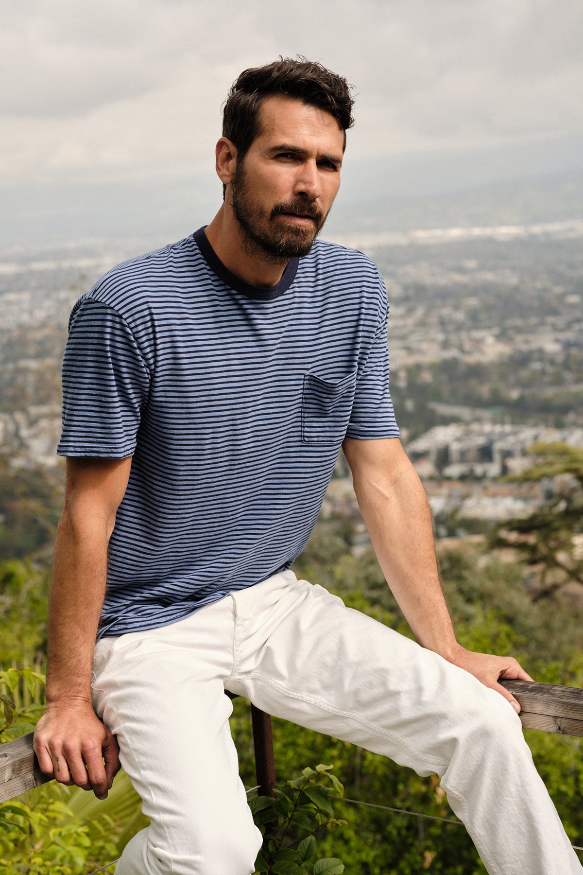   Lifestyle image of male model sitting on wooden railing overlooking a valley wearing Jeremy Crew Neck Tee with medium and dark blue stripes, front pocket, paired with white denim 