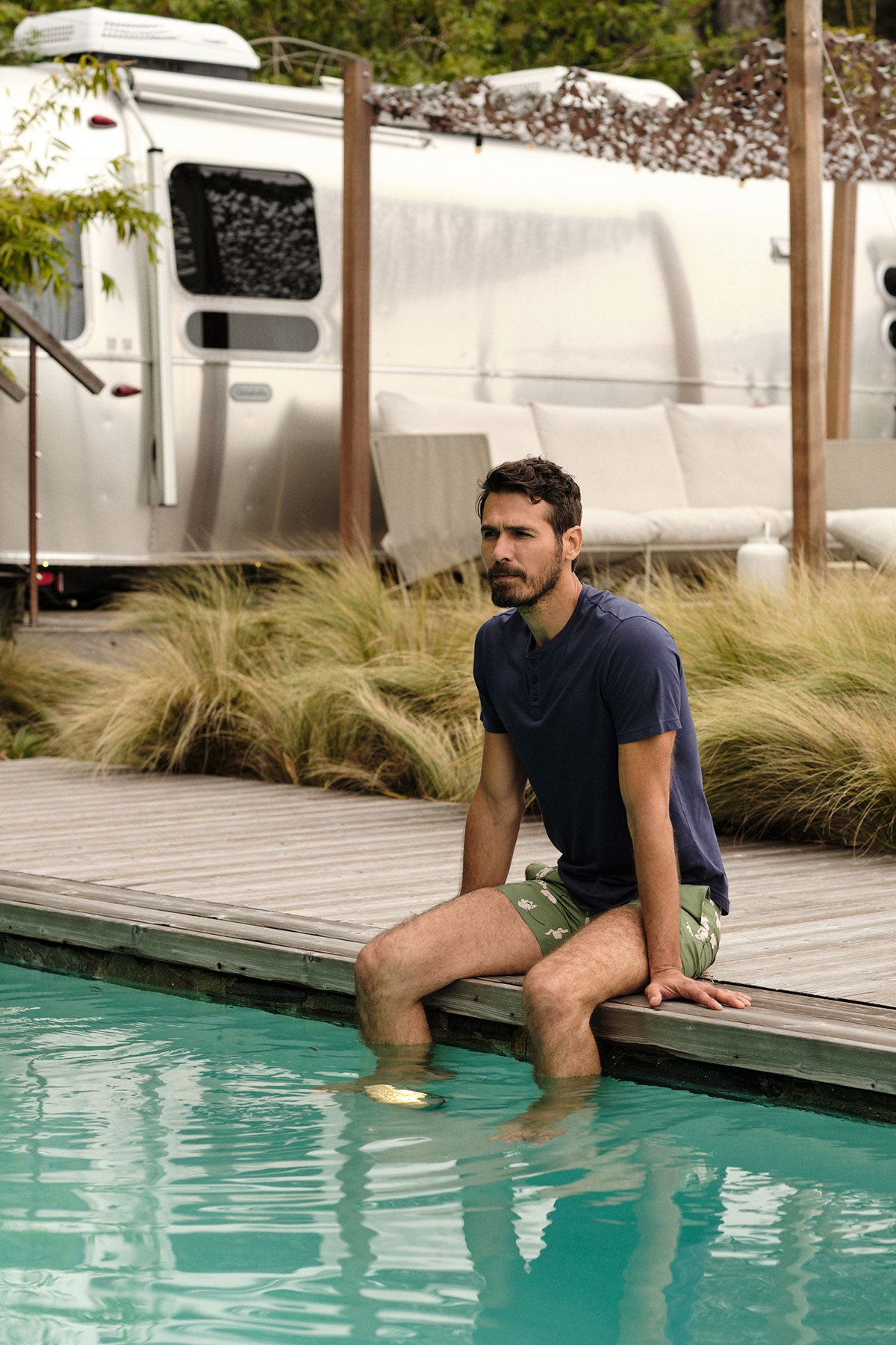   Lifestyle image of model sitting on edge of pool with feet in water wearing Fulton Short Sleeve Henley in midnight Ricardo Swim Short in green with white floral, Airstream trailer in the background 