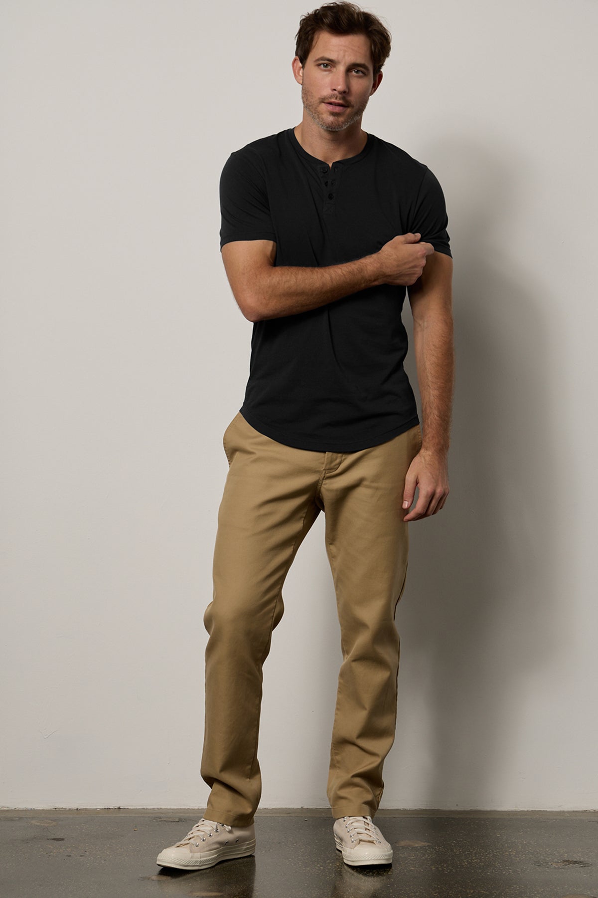  Man in black Fulton Henley and beige pants standing with one arm crossed over his chest, against a gray background. 