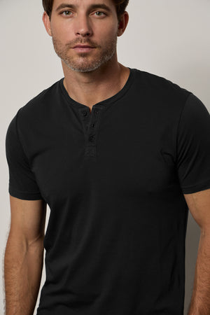 A man wearing a black Velvet by Graham & Spencer Fulton Henley tee, looking to the side, with a neutral expression on a plain background.