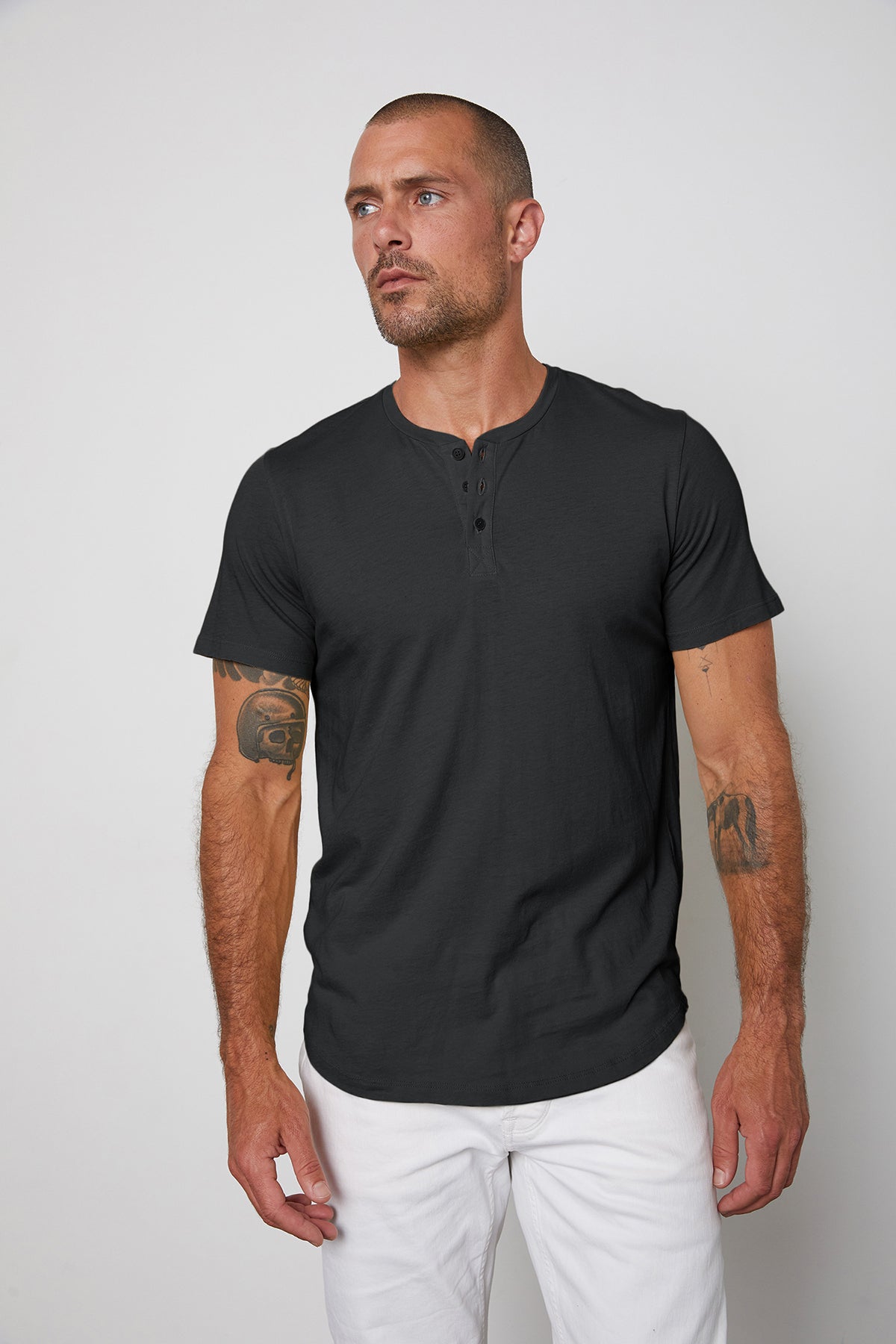 A man with a shaved head and tattoos on his arms wearing a black Velvet by Graham & Spencer Fulton Henley tee and white pants against a plain background.-24559747399873