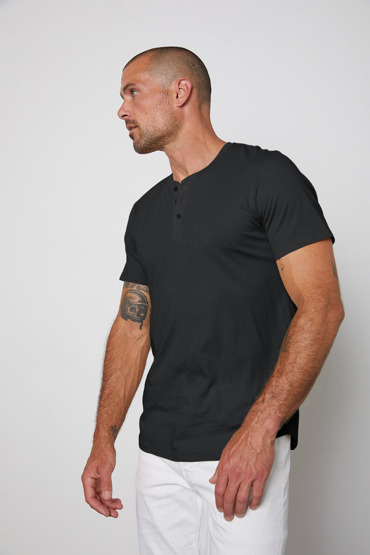   A man in a black Velvet by Graham & Spencer Fulton Henley and white pants, looking to the side, displaying a tattoo on his left arm against a plain background. 