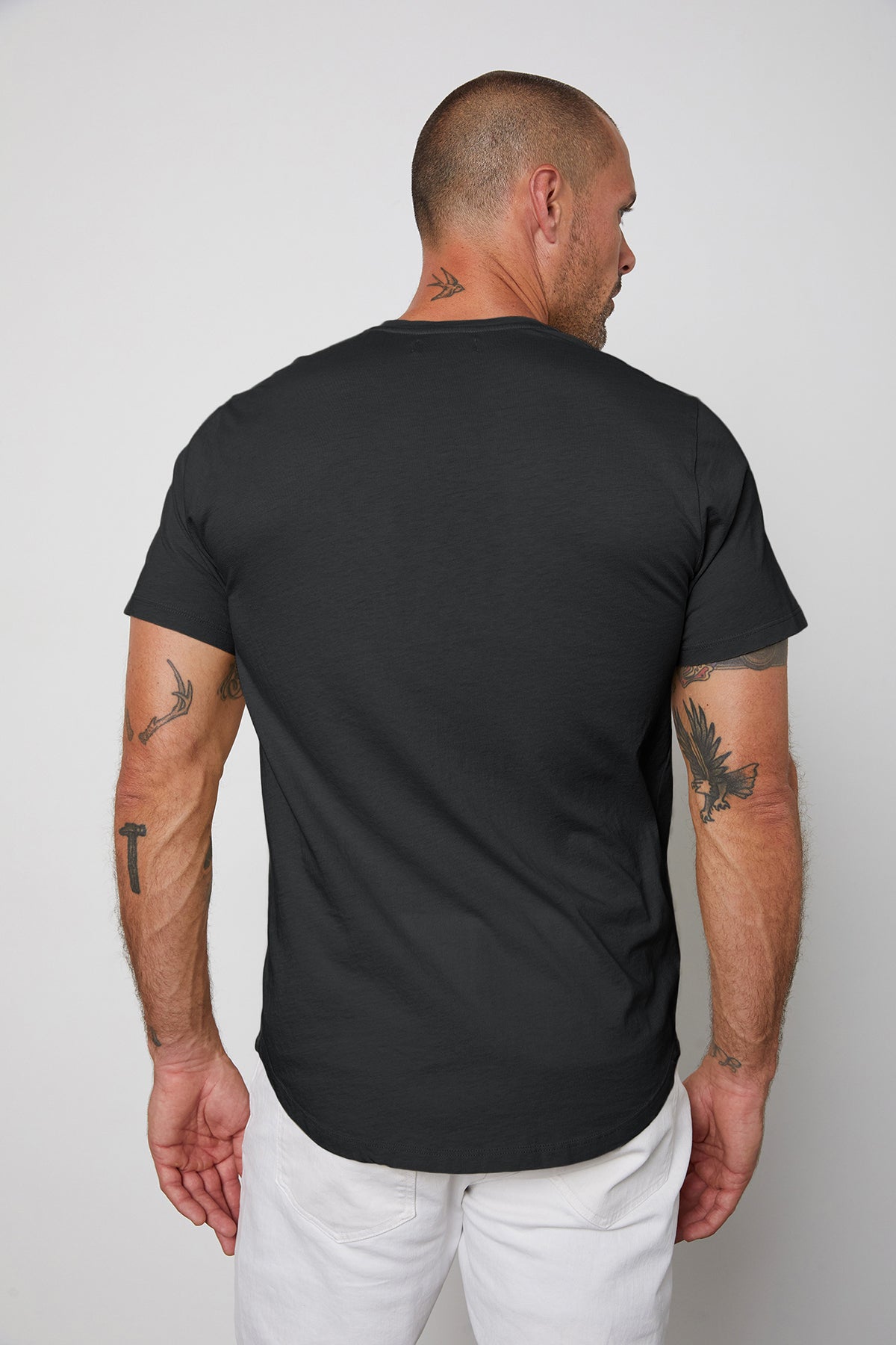 Man standing with his back to the camera, wearing a black Velvet by Graham & Spencer Fulton Henley and white pants, displaying tattoos on his arms.-24559747465409