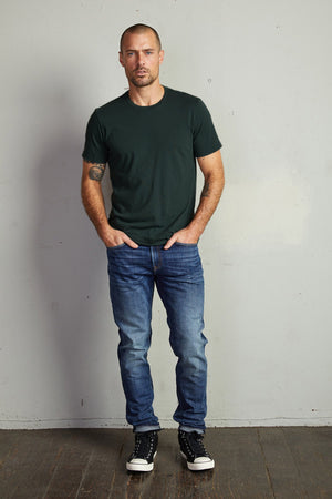 A man with a shaved head is standing against a plain wall, wearing a dark green HOWARD TEE by Velvet by Graham & Spencer with a vintage feel, blue jeans, and black sneakers. He has his hands in his pockets and a tattoo on his left arm. The HOWARD TEE, made of lightweight cotton knit, looks like the perfect fit for him.