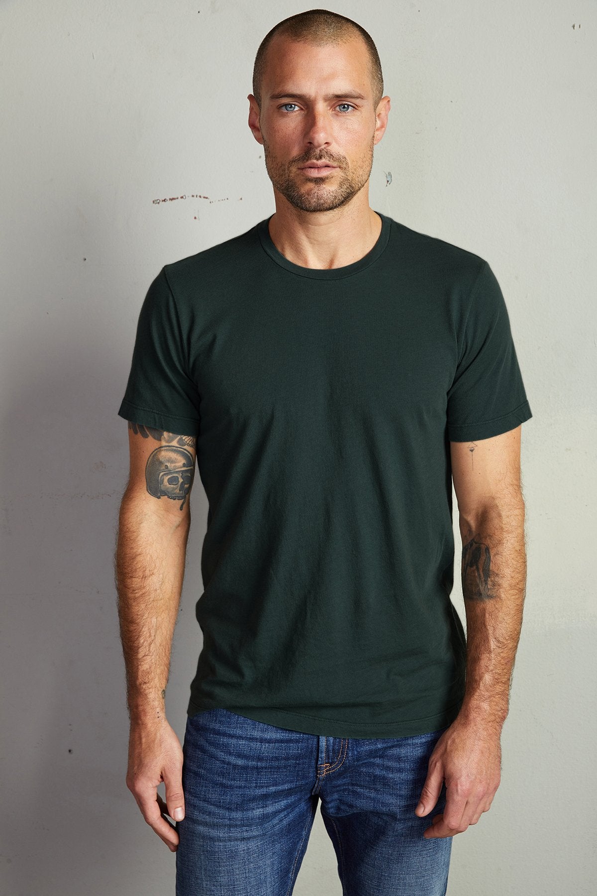   A man with short hair and tattoos on both forearms stands against a plain wall, wearing a dark green Velvet by Graham & Spencer HOWARD TEE with a perfect fit and blue jeans. 