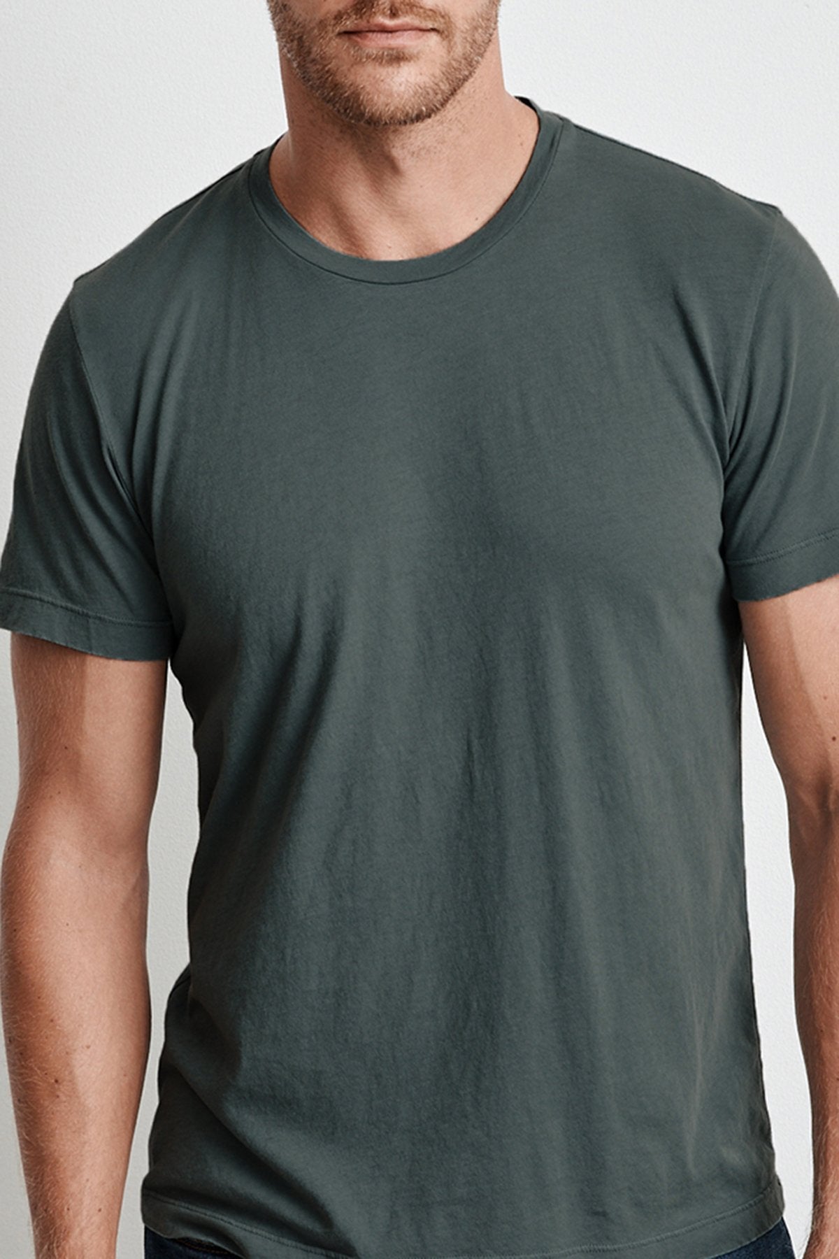   A man wearing a plain dark green HOWARD TEE by Velvet by Graham & Spencer, made from lightweight cotton knit, stands with his arms at his sides, facing the camera. The background is a neutral light color. Only his upper body is visible. 