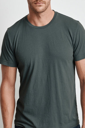 A man wearing a plain dark green HOWARD TEE by Velvet by Graham & Spencer, made from lightweight cotton knit, stands with his arms at his sides, facing the camera. The background is a neutral light color. Only his upper body is visible.