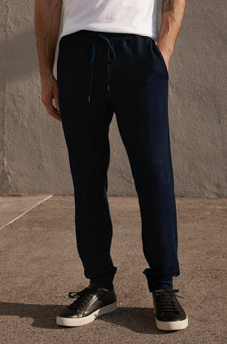 A person wearing a white t-shirt, Velvet by Graham & Spencer CROSBY LUXE FLEECE JOGGER pants, and black sneakers with white soles stands on a paved area in front of a gray wall, possibly getting ready for their workouts.-25051978367169