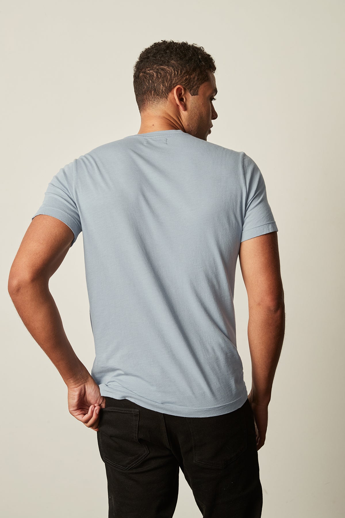   A man with short hair stands with his back to the camera, wearing a light blue T-shirt with a subtle v-neckline and black pants, showcasing the airy comfort of the SAMSEN WHISPER CLASSIC V-NECK TEE by Velvet by Graham & Spencer. 