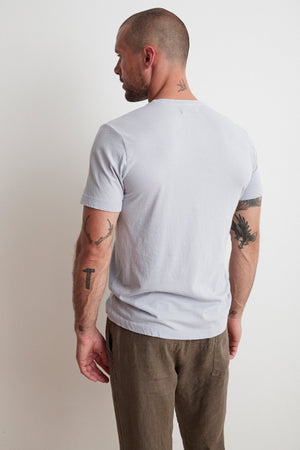 A man with a shaved head and tattoos on his arms and neck stands facing away, wearing a light gray SAMSEN WHISPER CLASSIC V-NECK TEE by Velvet by Graham & Spencer and brown pants, ideal for everyday wear, against a plain white background.