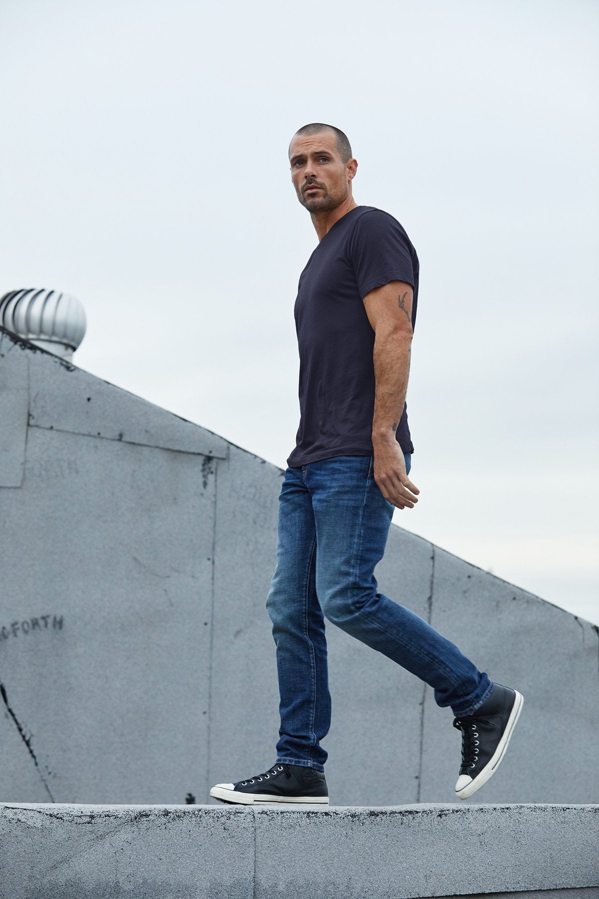   A man wearing a dark SAMSEN WHISPER CLASSIC V-NECK TEE by Velvet by Graham & Spencer, jeans, and black sneakers is walking on a concrete surface. A triangular structure and some industrial elements are visible in the background. 