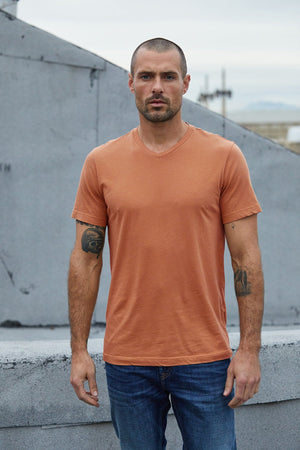 A man with short hair and tattoos on both arms stands outdoors wearing an orange SAMSEN WHISPER CLASSIC V-NECK TEE by Velvet by Graham & Spencer, paired with blue jeans, perfect for everyday wear.