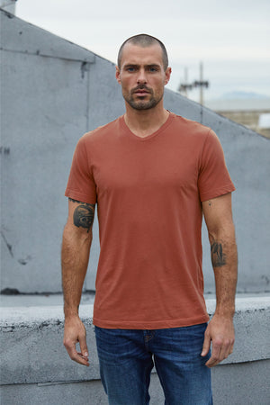 A man with close-cropped hair and tattoos on his arms stands outdoors wearing a rust-colored Velvet by Graham & Spencer SAMSEN WHISPER CLASSIC V-NECK TEE and blue jeans.