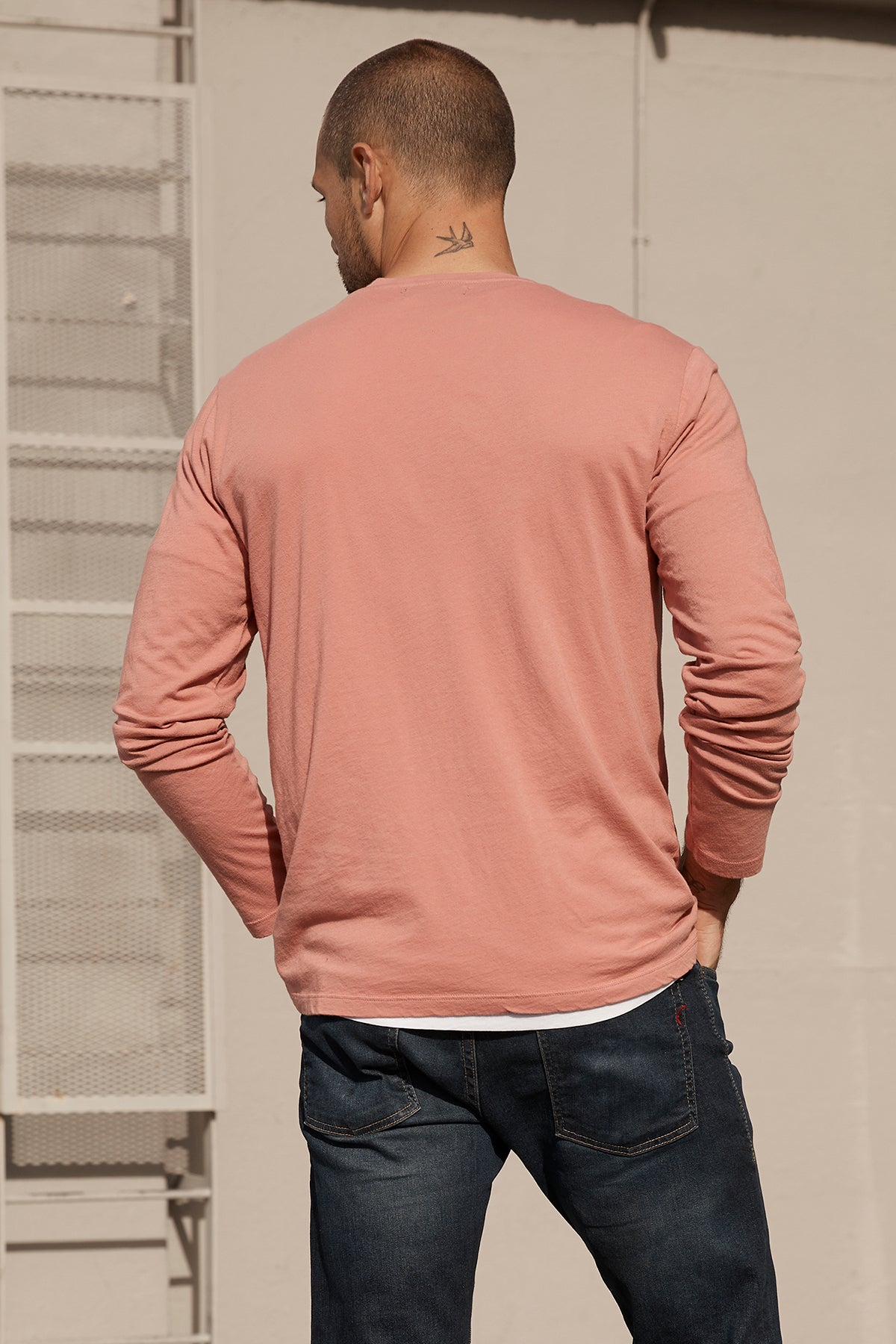   Back view of a person with short hair wearing an ultra-soft cotton long-sleeve pink Velvet by Graham & Spencer SKEETER WHISPER CLASSIC CREW NECK TEE and dark jeans, standing outdoors. 