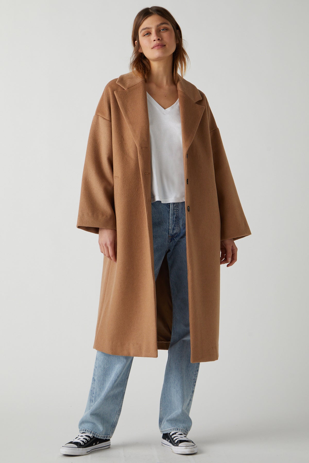 A person stands wearing a classic HUNTINGTON COAT by Velvet by Jenny Graham over a white V-neck top, blue jeans, and black and white sneakers. The chic essential outfit is set against a plain, light-colored background.-25483423514817