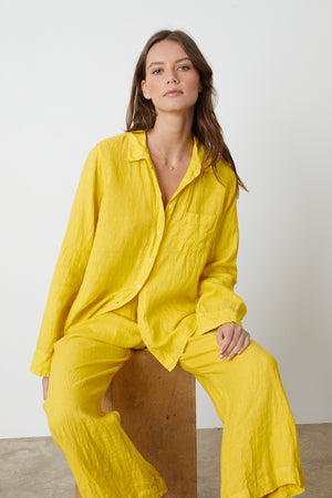Model sitting on wooden box stool wearing Natalia Button-Up Shirt in bright yellow sun colored linen with Lola pant front