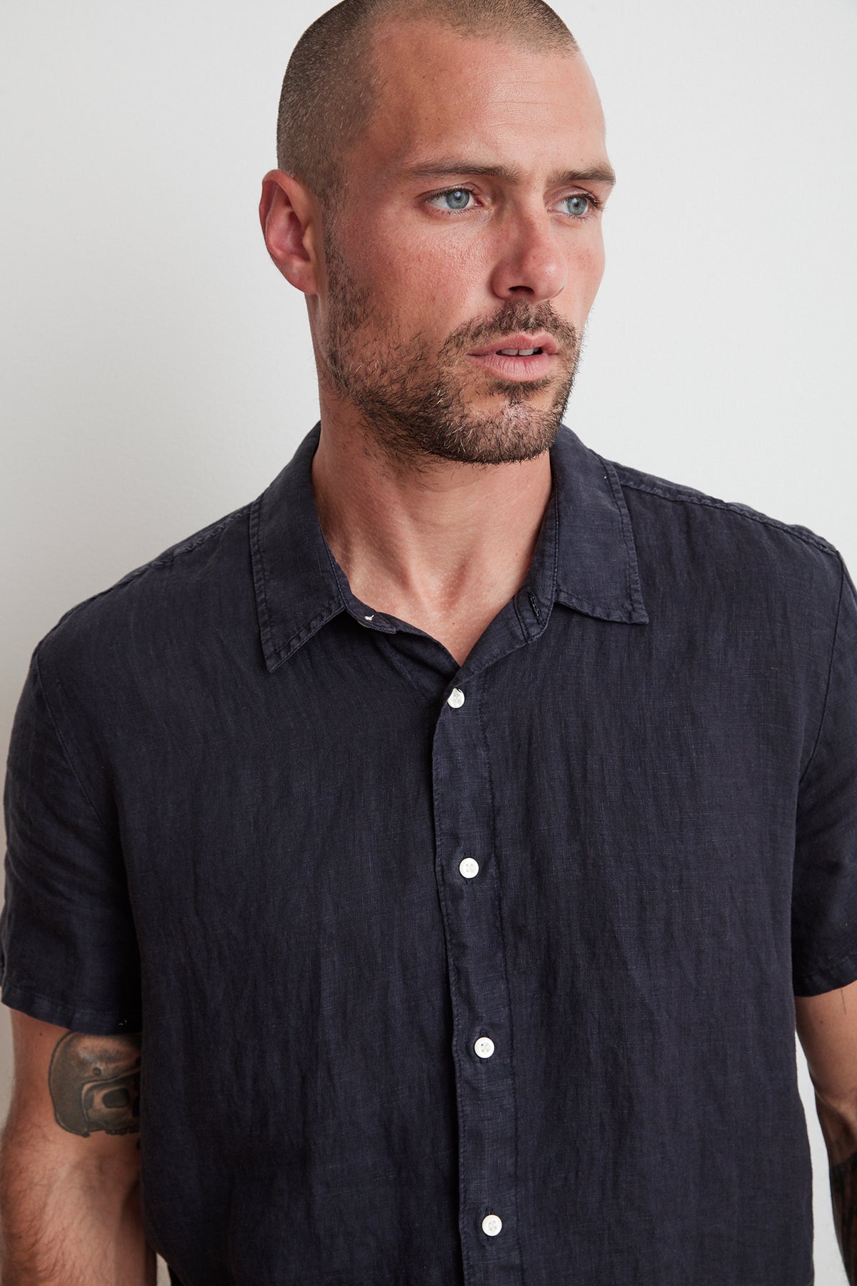   A man with a shaved head, beard, and tattoo on his left arm is wearing a short-sleeved, dark-colored MACKIE LINEN BUTTON-UP SHIRT by Velvet by Graham & Spencer while looking slightly to his left. 