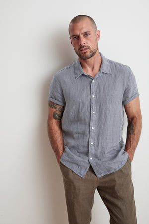 A man with a shaved head and beard, wearing a short-sleeved, relaxed fit grey MACKIE LINEN BUTTON-UP SHIRT by Velvet by Graham & Spencer and brown pants, stands against a plain white wall with his hands in his pockets.