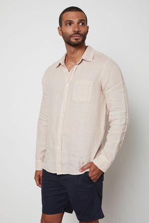 A man wearing a beige BENTON LINEN BUTTON-UP SHIRT by Velvet by Graham & Spencer and dark shorts stands against a plain white background, looking to his right.