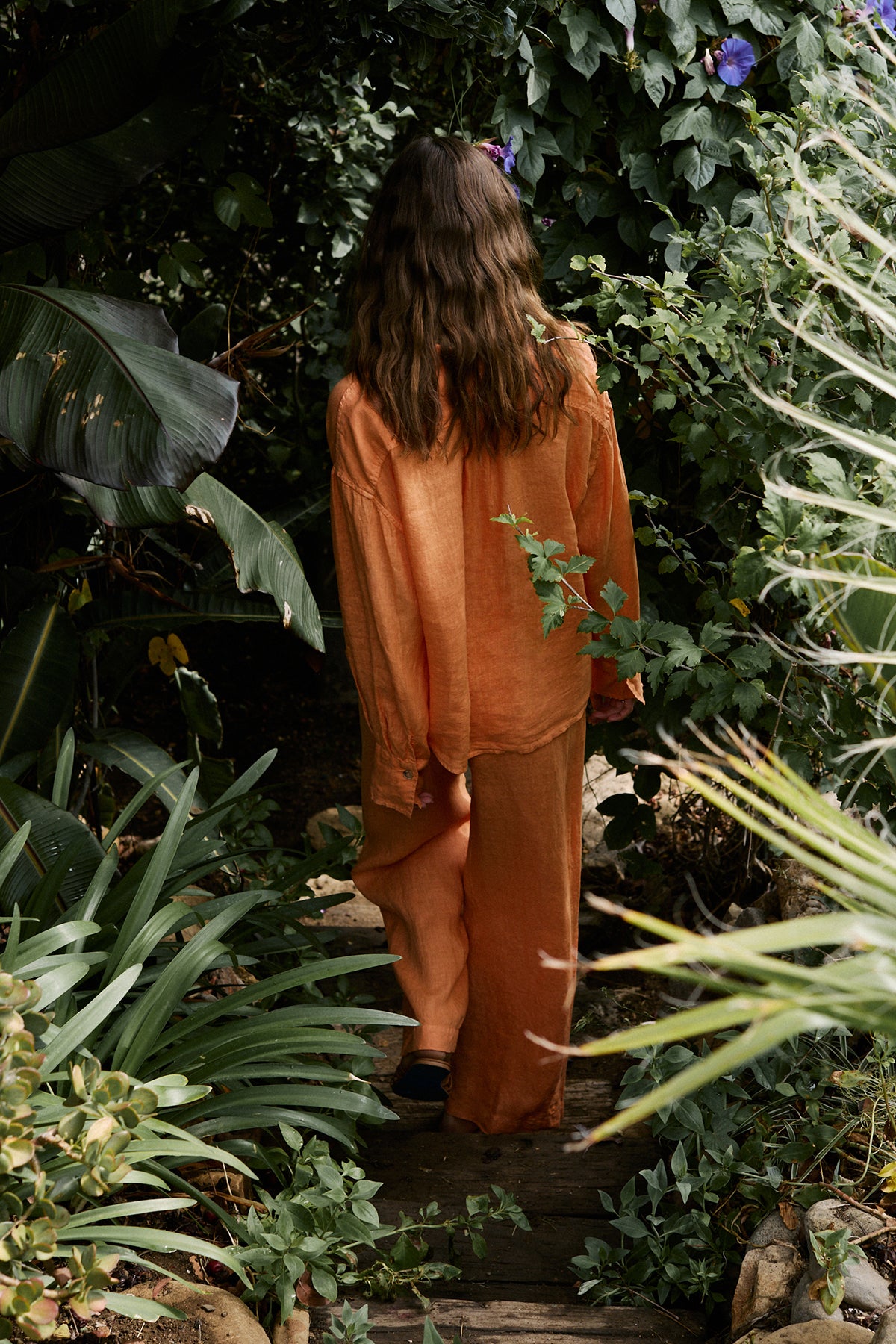   Women walking down wooden steps outdoors surrounded by trees wearing Eden shirt in orange heat with Lola pant, necklaces and sandals full length back 