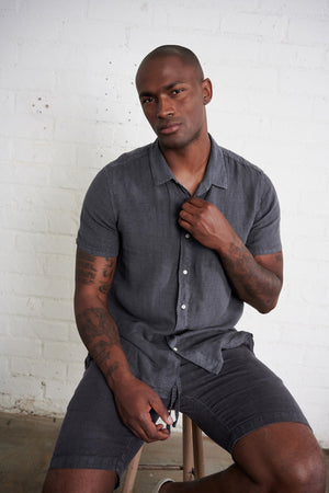 Mackie linen button up shirt in carbon with model sitting on stool.
