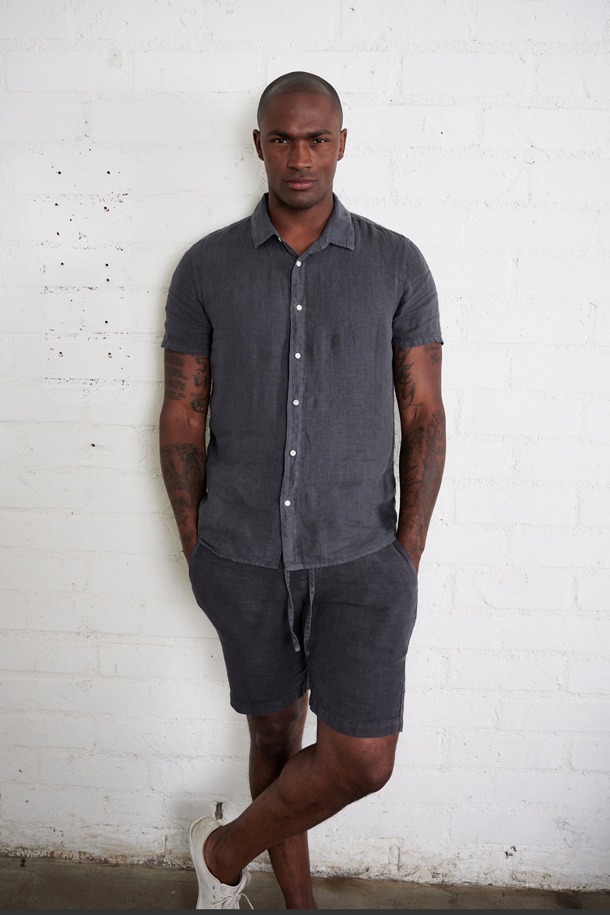   Mackie linen button up shirt in carbon with model standing against white brick wall with hands in pockets. 