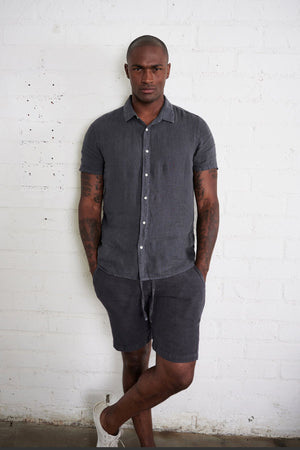 Mackie linen button up shirt in carbon with model standing against white brick wall with hands in pockets.