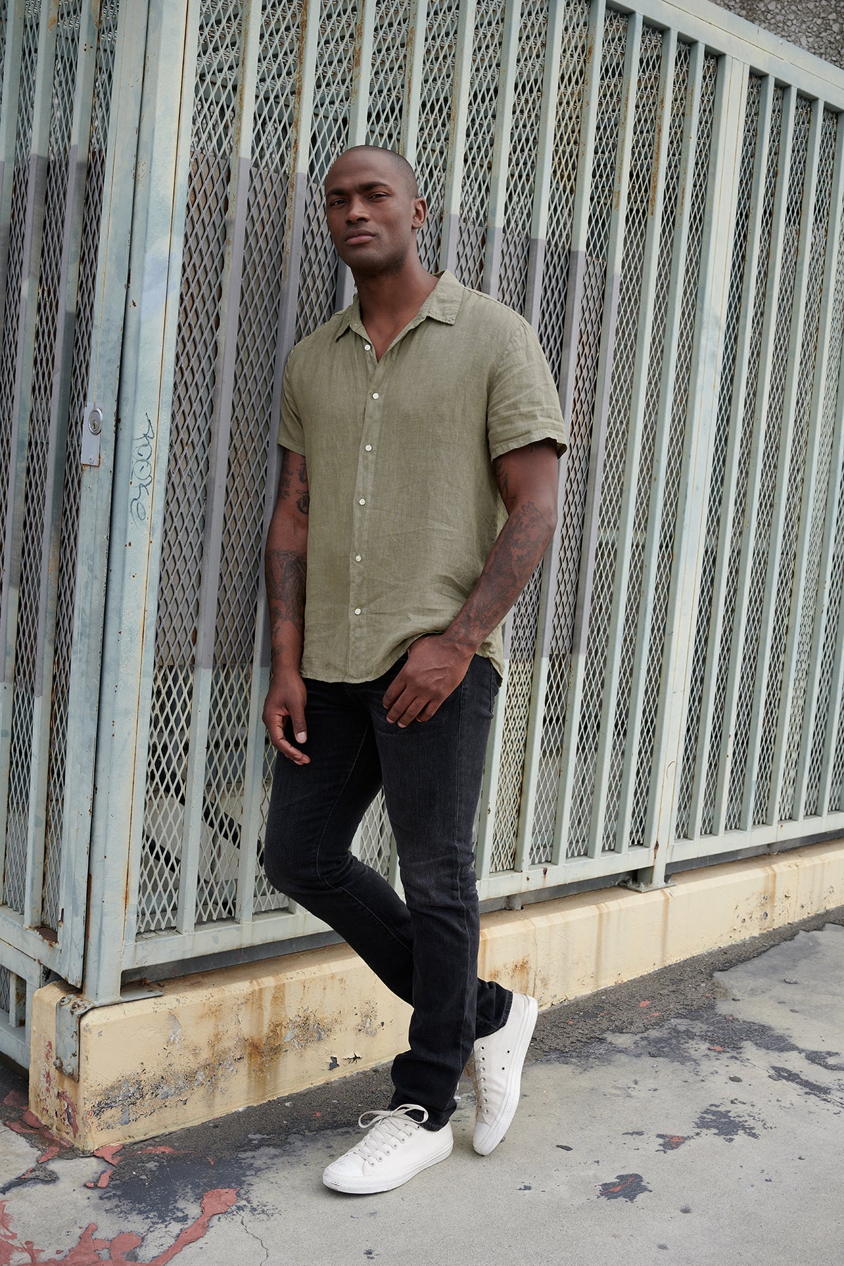   Mackie linen button up shirt in olive with black denim and white sneakers with model leaning against metal fence. 