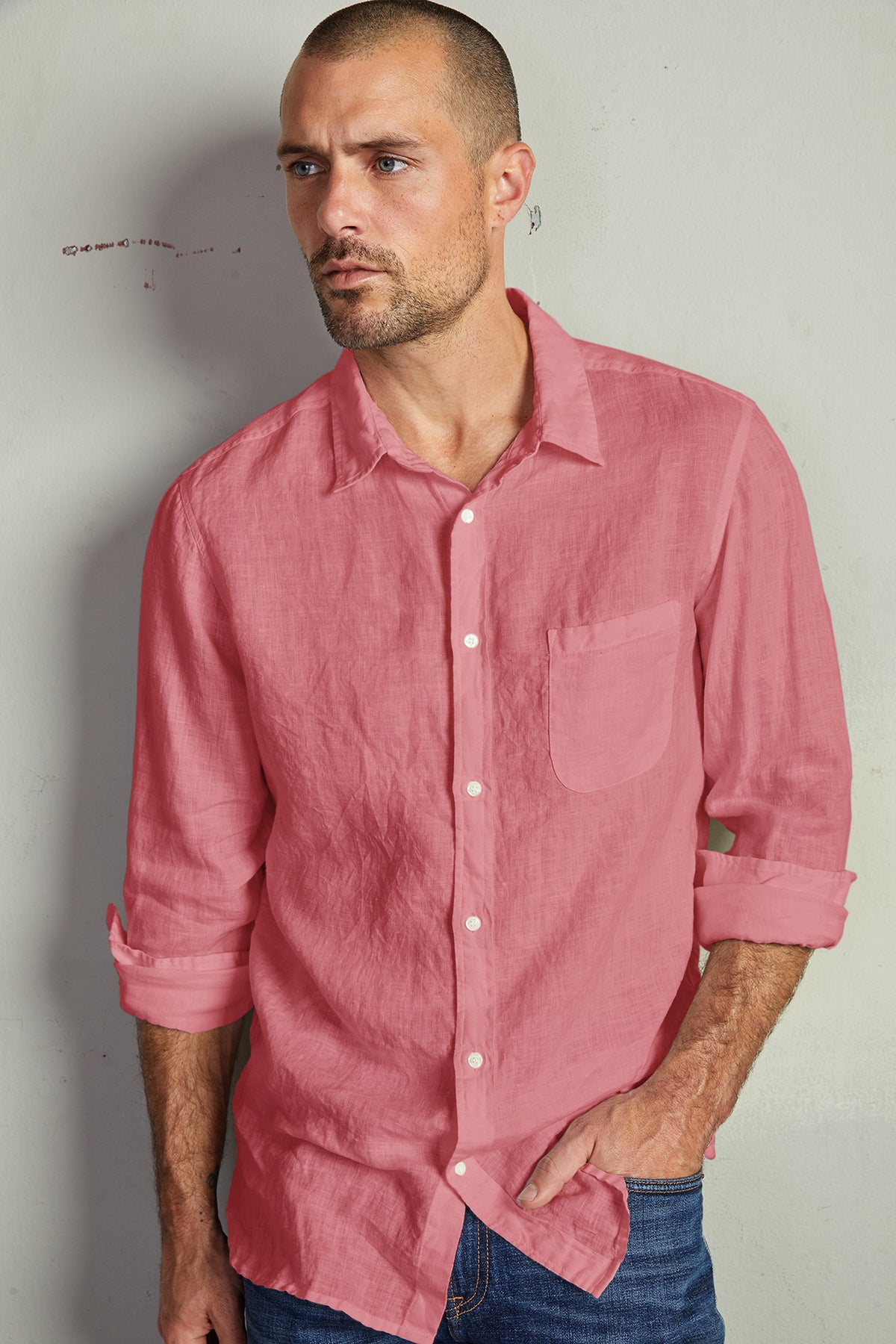   A man with a shaved head and beard, wearing the BENTON LINEN BUTTON-UP SHIRT by Velvet by Graham & Spencer in pink, paired with blue jeans, stands against a plain background. 