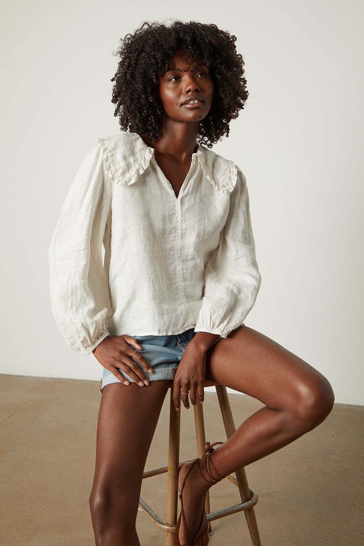 Model sitting on stool in studio wearing Sofia linen top in beach with blue denim shorts front-26079070290113