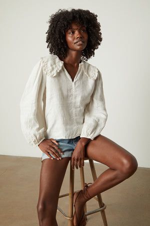 Model sitting on stool in studio wearing Sofia linen top in beach with blue denim shorts front