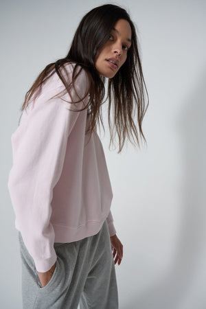 A person with long dark hair wearing a pink YNEZ SWEATSHIRT by Velvet by Jenny Graham and gray pants is standing and looking slightly to the side.