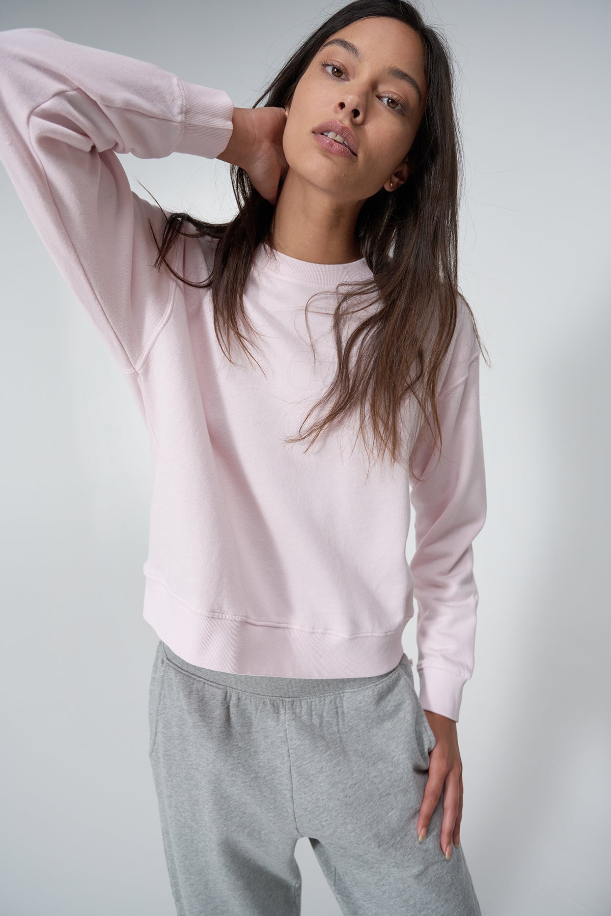   A person with long hair is standing and posing with one hand behind their head. They are wearing a light pink YNEZ SWEATSHIRT by Velvet by Jenny Graham and gray pants. 